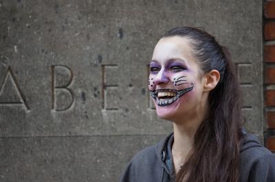 Lachende Frau mit breiter geschminktem Mund, vor einer Betonwand mit eingelassenen Buchstaben (sichtbar nur &quot;A&quot;, &quot;B&quot; und &quot;E&quot;.