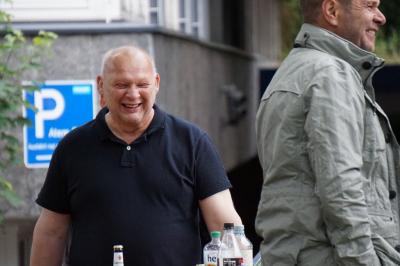 Lachender, etwa 45jähriger Mann in blauem T-Shirt mit Halbglatze, im Vordergrund ein weiterer lachender Mann von der Seite (in beiger Jacke), im Hintergrund ein Gebäude und ein &quot;Parkplatz&quot;-Schild