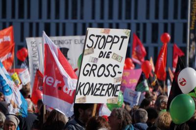 Menschen, Fahnen, dazu ein Papp-Plakat mit der Aufschrift &quot;Stoppt die Gross-Konzerne&quot;.