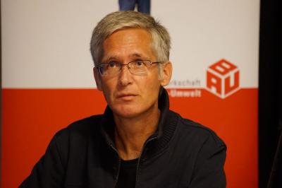 Grauhaariger Mann dunklem Teint und Brille, im Hintergrund der &quot;BAU&quot;-Würfel der IG BAU.