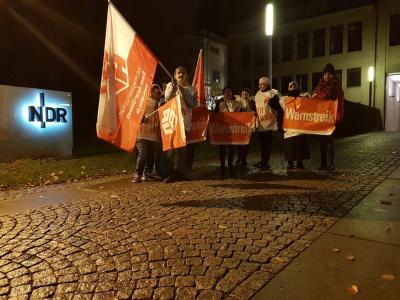Etwa ein Dutzend Menschen, überwiegend mit rot-weißen Streikplakaten und Gewerkschaftsfahnen auf einer gepflasterten Auffahrt. Im Hintergrund der beleuchtete Schriftzug &quot;NDR&quot;. Es ist noch dunkel, im Hintergrund leuchtet eine Straßenlaterne.