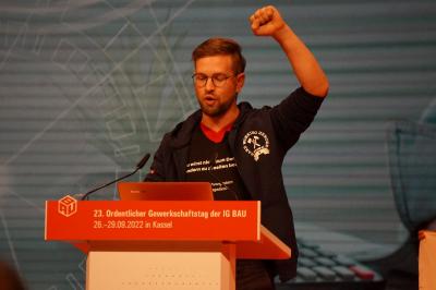 Redner "Cirka" von der Jungen BAU mit erhobener linker Faust am Rednerpult des Gewerkschaftstages Redner "Cirka" von der Jungen BAU mit erhobener linker Faust am Rednerpult des Gewerkschaftstages