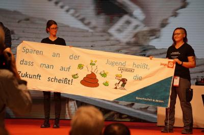 Zwei junge Gewerkschafterinnen mit einem Transparent auf der Bühne. Darauf abgebildet u.a. ein Haufen Scheiße und der Slogan: Sparen an der Jugend heißt, dass man auf die Zukunft scheißt. Zwei junge Gewerkschafterinnen mit einem Transparent auf der Bühne. Darauf abgebildet u.a. ein Haufen Scheiße und der Slogan: Sparen an der Jugend heißt, dass man auf die Zukunft scheißt.
