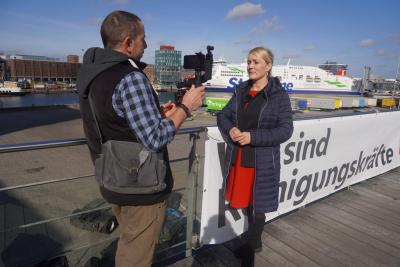 Eine Frau wird vor einem Hafenbecken interviewt, ein Journalist filmt sie mit einer Handykamera. Im Hintergrund ist ein Kreuzfahrtschiff der Stena Line und ein Teil des Kieler Hafens zu sehen.