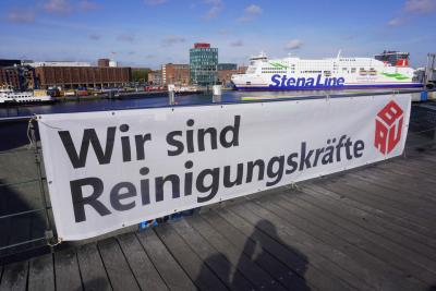 Banner mit der Aufschrift "Wir sind Reinigungskräfte" (schwarz auf weiß) und BAU-Logo, im Hintergrund ein Hafenbecken mit einem Kreuzfahrtschiff der Stena-Line.