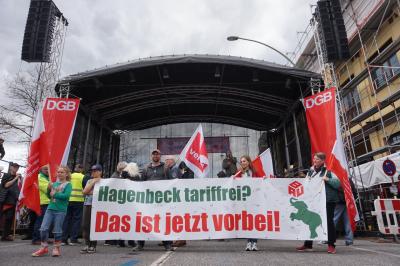 Etwa ein dutzend Menschen vor ver.di-Fahnen und DGB-Bannern tragen ein Transparent mit der Aufschrift "Hagenbeck tariffrei? Das ist jetzt vorbei!". Im Hintergrund eine große Bühne. Etwa ein dutzend Menschen vor ver.di-Fahnen und DGB-Bannern tragen ein Transparent mit der Aufschrift "Hagenbeck tariffrei? Das ist jetzt vorbei!". Im Hintergrund eine große Bühne.