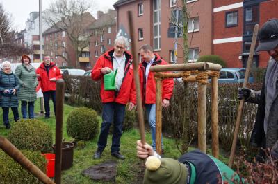 Mehrere Männer in roten IG BAU-Jacken, einer von ihnen mit einer grünen Gießkanne. Im Vordergrund ein frischgepflanzter Baum in einem stützenden Gerüst. Mehrere Männer in roten IG BAU-Jacken, einer von ihnen mit einer grünen Gießkanne. Im Vordergrund ein frischgepflanzter Baum in einem stützenden Gerüst.