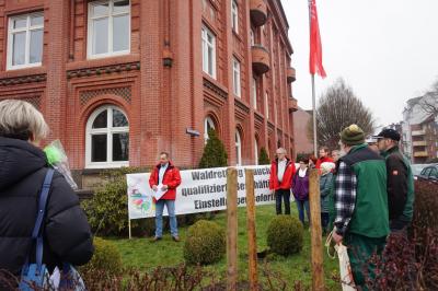 Ein Mann in roter IG BAU-Jacke spricht vor einem Transparent im Garten eines roten Backsteinhauses. Knapp ein dutzend Menschen hören zu. Ein Mann in roter IG BAU-Jacke spricht vor einem Transparent im Garten eines roten Backsteinhauses. Knapp ein dutzend Menschen hören zu.