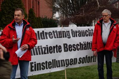 Zwei Männer in roten IG BAU-Jacken vor einem Transparent. Schwarze Schrift auf weißem Stoff: "Waldrettung braucht qualifizierte Beschäftigte - Einstellungen sofort!" Zwei Männer in roten IG BAU-Jacken vor einem Transparent. Schwarze Schrift auf weißem Stoff: "Waldrettung braucht qualifizierte Beschäftigte - Einstellungen sofort!"