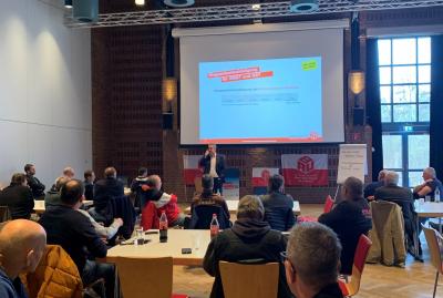 Bild einer Veranstaltung mit etlichen sitzenden Menschen, die zu einem Redner und inr großen Leinwand schauen. Im Hintergrund rechts ein Fenster, in das die Sonne scheint. Bild einer Veranstaltung mit etlichen sitzenden Menschen, die zu einem Redner und inr großen Leinwand schauen. Im Hintergrund rechts ein Fenster, in das die Sonne scheint.